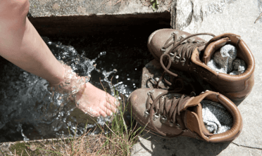 With their removed hiking boot off to one side, walker washes their foot in clear water.
